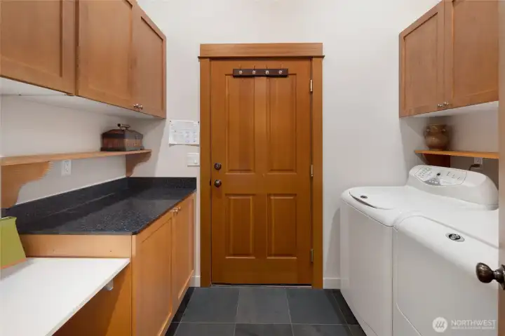 The well-designed laundry room impresses with warm wood cabinetry on both sides, dark granite countertop workspace, and slate tile flooring for a polished, functional finish. A solid wood interior door to garage and full-size washer & dryer complete this thoughtfully appointed utility space.
