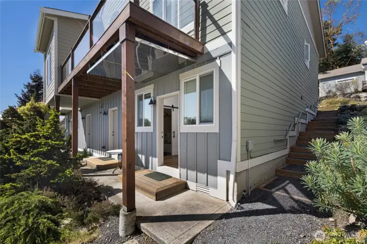 Charming lower-level entry to the separate studio with fill bath features a covered porch with warm wood beam accents and lush mature landscaping, offering a welcoming and private first impression. The modern board-and-batten siding in soft gray tones complements the natural surroundings of San Juan Island.
