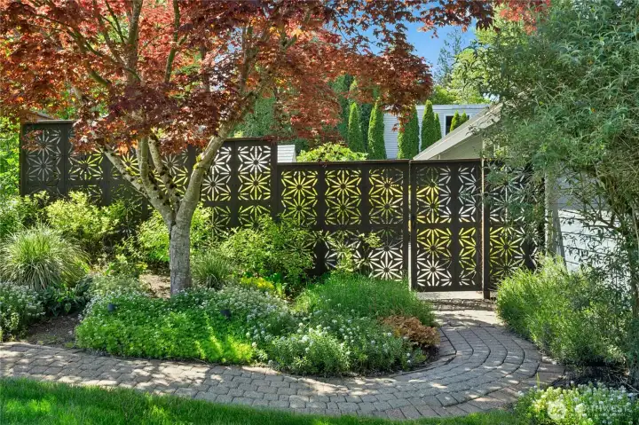 Custom decorative fencing adds both privacy and artistic character to the landscape, creating a striking backdrop framed by mature trees and lush garden beds.  Curved pathways and layered greenery enhance the peaceful, thoughtfully designed outdoor setting.
