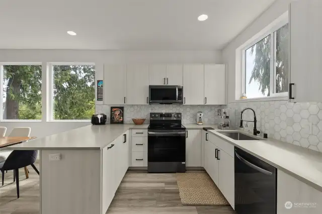 This bright, modern kitchen boasts sleek black stainless steel appliances and custom cabinetry paired with a striking hexagonal tile backsplash. Ample natural light flows through large windows, illuminating the space and providing serene views of the surrounding trees.