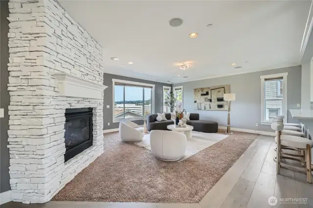 This beautifully staged living room features a modern design with a striking white stone fireplace as the focal point. Large windows fill the space with natural light and offer scenic views, while the soft gray walls and plush carpet create a warm and inviting atmosphere. Stylish furniture, including a curved sectional and two contemporary swivel chairs, is arranged around a chic round coffee table, perfectly balancing comfort and elegance.