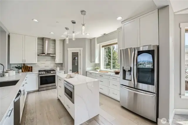 Chef-approved and magazine-worthy, this light-filled kitchen pairs crisp contemporary styling with everyday practicality. White shaker cabinetry stretches to the ceiling, offering abundant storage and framing a sleek stainless-steel appliance suite—including a gas range with designer hood and a French-door fridge with InstaView window. Veined quartz countertops flow seamlessly into a waterfall-edge island that doubles as a casual breakfast spot and houses a built-in microwave for clutter-free counters. A soft-toned mosaic backsplash, trio of glass-pendant lights, and wide-plank hardwood floors add layers of texture and warmth, while the walk-in pantry with etched-glass door keeps essentials neatly organized. Whether you’re hosting a dinner party or packing school lunches, this thoughtfully planned culinary hub makes every task feel effortlessly elegant.