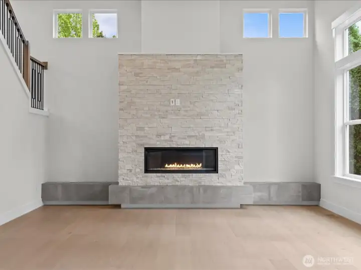 A closer look at the living area showcasing high ceilings, rich wide-plank engineered hardwood floors, and a dramatic stone fireplace surround featuring a functional raised hearth designed for extra seating.