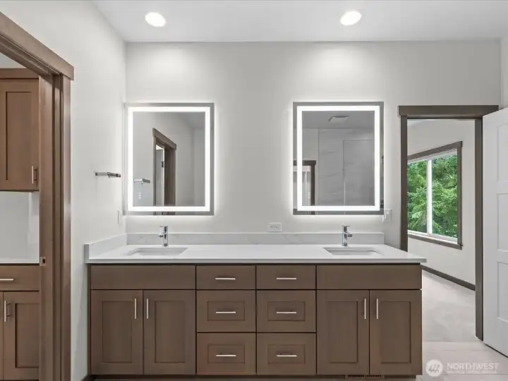 Gorgeous primary bathroom double vanity featuring rich wood cabinetry, premium quartz countertops, and contemporary undermount sinks. Perfectly accented by dual backlit LED mirrors and bright, modern fixtures.