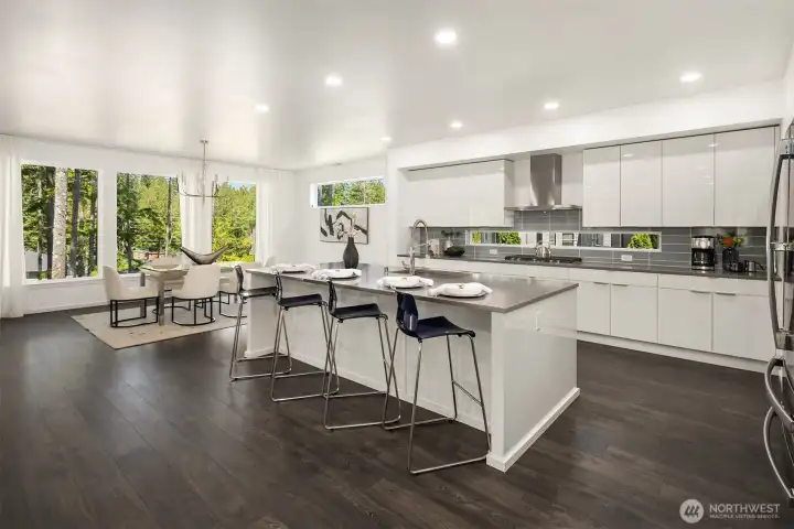 Open-concept kitchen and dining area with sleek modern finishes, oversized island seating, abundant natural light, and views toward the mature greenbelt.