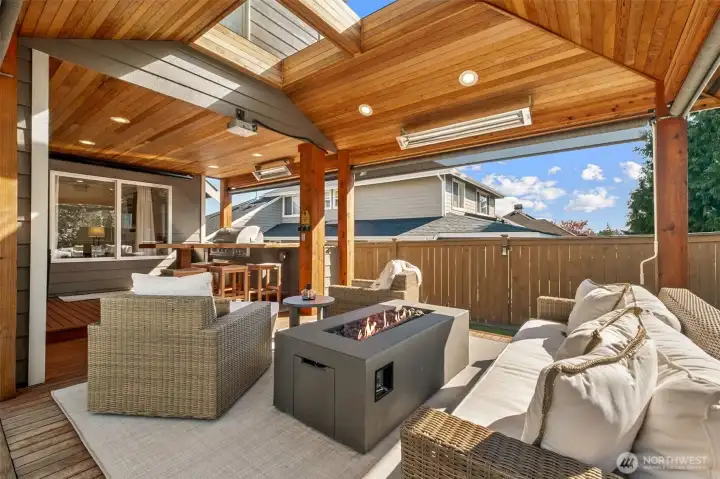 Back to stepping out through those French Doors off the kitchen ... Resort-style covered outdoor living addition showcasing a vaulted wood ceiling with skylights, integrated heaters, a modern gas fire table, and a built-in grilling area—creating a seamless, year-round extension of the home.