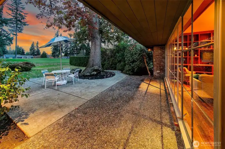 A covered walkway extends along the glass façade toward the bungalow, opening to an intimate patio framed by mature plantings and the manicured fairway beyond. This sheltered outdoor zone creates a fluid connection between indoor living and the course-side landscape, offering a calm pause before stepping into the home’s freestanding bungalow wing.