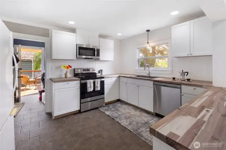 The fully updated kitchen showcases stainless steel appliances including a range, over-the-range microwave, and dishwasher, complemented by warm butcher block counters and ample white cabinetry. A pendant light and shiplap accent wall add charm, while a doorway connects easily to the entry and deck for effortless indoor-outdoor living.
