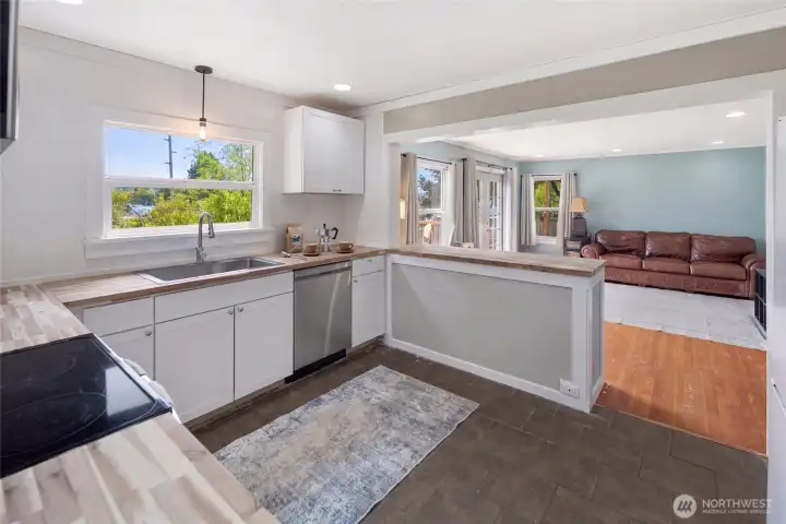 The freshly renovated kitchen features crisp white shaker cabinets, butcher block countertops, stainless steel dishwasher, and a peninsula that opens directly to the living room — ideal for entertaining. White shiplap-style walls and a large window flood the space with natural light and a bright, coastal aesthetic.