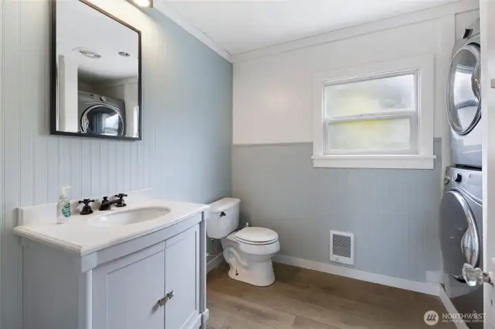 The tastefully remodeled bathroom features beadboard wainscoting in calming blue-gray tones, tiled shower, a white shaker vanity, and the added convenience of a stacked washer and dryer — combining style and everyday functionality in one well-appointed space.