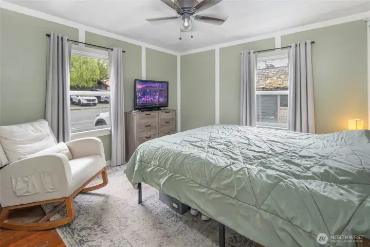 The sun-filled bedroom showcases three windows framing leafy neighborhood views, a ceiling fan, and tasteful sage-green walls with white board-and-batten detail that echo the home's refreshed, cohesive design.