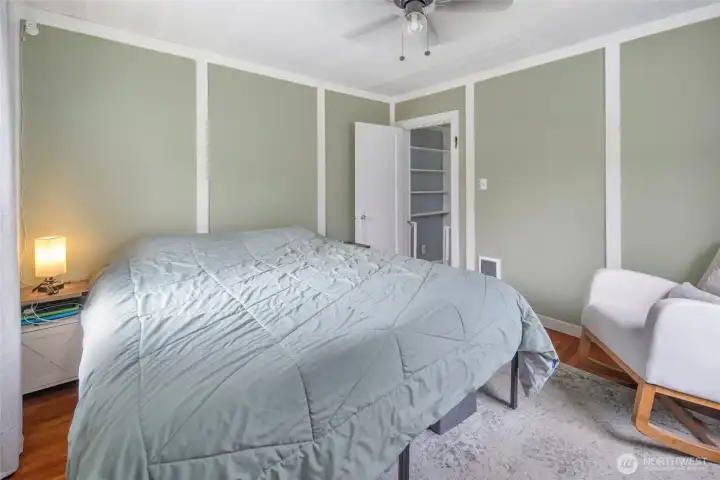 This inviting secondary bedroom showcases a charming board-and-batten wall treatment in soft sage green, complemented by warm hardwood floors, a ceiling fan, and a cozy reading nook with accent chair — a restful retreat that reflects the cohesive, thoughtfully updated aesthetic found throughout the home.