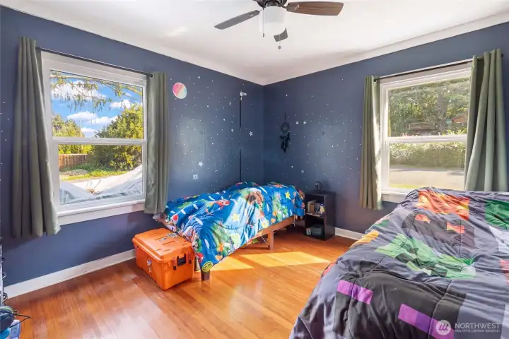 A charming bedroom with warm hardwood floors features two large windows that frame neighborhood views and fill the room with natural light, complemented by a ceiling fan.