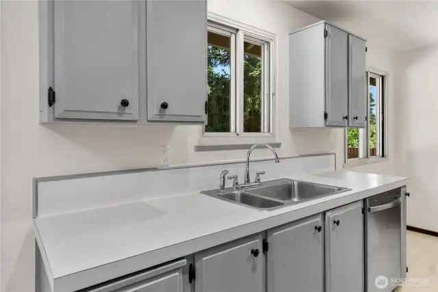 Bright galley kitchen with freshly painted cabinetry and a double-basin sink set beneath dual windows. The upgraded quadruple-pane windows provide natural light while adding energy efficiency and soundproofing.