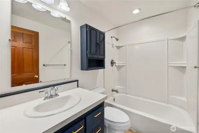 The home’s full bathroom offers a clean, functional design with a shower/tub combo featuring built-in shelving, a bright vanity with storage, and overhead lighting. Bold navy cabinetry with gold hardware provides a stylish contrast, while the neutral finishes keep the space fresh and easy to personalize.