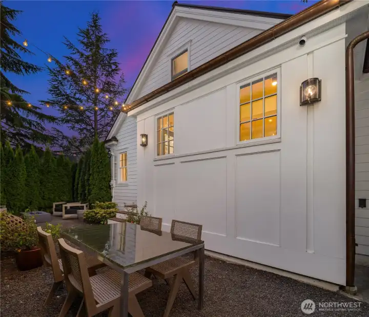 An elegant garden dining area, framed by lush evergreens and glowing café lights, offers the perfect setting for al fresco meals long into the evening.