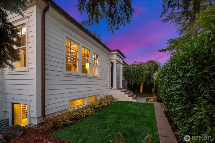 The front elevation glows at dusk, where crisp architectural lines meet a carefully curated, serene and evergreen garden approach for privacy.