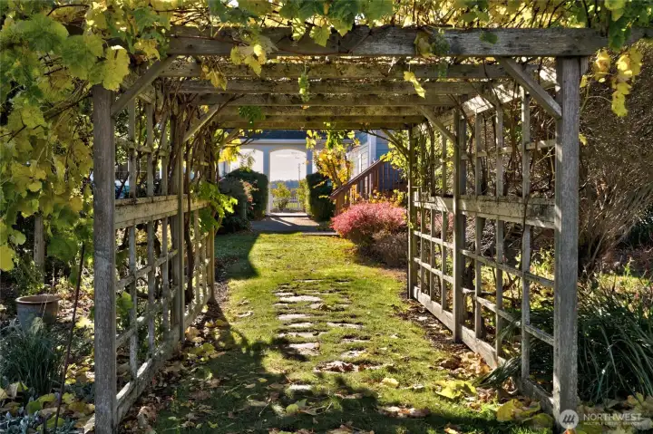 A beautiful trellis of purple wisteria, red roses, and grapevines connects to the fenced vegetable gardens and a large glass greenhouse.