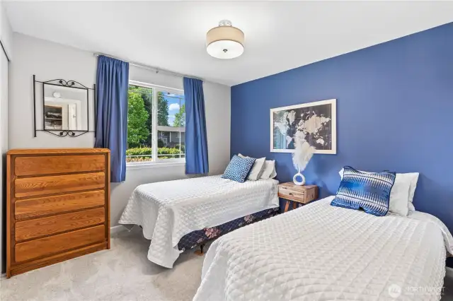 Charming and light-filled guest bedroom with dual twin beds, a bold navy accent wall, large picture window, and plush carpeting. Enjoy serene front yard views and ample natural light in this beautifully staged space—perfect as a guest suite or home office.
