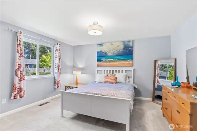 Expansive primary bedroom with soft natural light, calming gray walls, and plush carpeting. Features a spacious layout, large window with private backyard views, and stylish touches including bold drapery and coastal-themed décor. A perfect retreat to unwind in comfort.