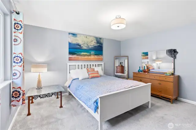 Relax in the serene primary bedroom featuring soft gray walls, plush carpet, and abundant natural light. The room offers generous space for a full furniture suite, including a large dresser and nightstands, with peaceful design elements that create a calm and cozy retreat.