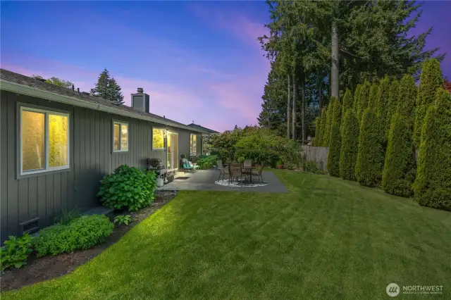 Enjoy peaceful evenings in this fully fenced backyard oasis featuring a spacious patio perfect for grilling, outdoor dining, and cozy gatherings. Surrounded by mature landscaping and tall evergreens for added privacy, this twilight view highlights the seamless indoor-outdoor lifestyle this home offers.