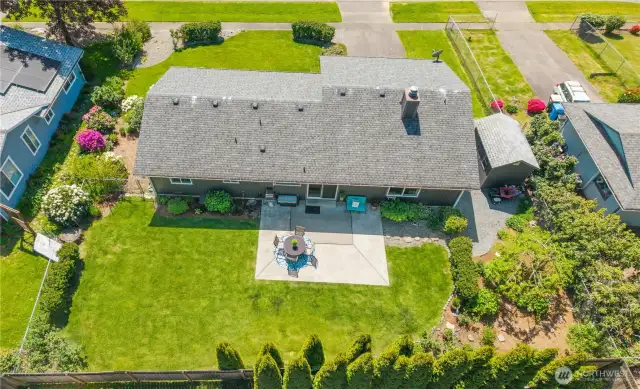 Stunning drone capture of the backyard oasis featuring a spacious concrete patio for outdoor dining, lush green lawn, and mature privacy landscaping. Thoughtfully designed with garden beds, flowering shrubs, and peaceful seating areas—perfect for entertaining or relaxing under the sun.