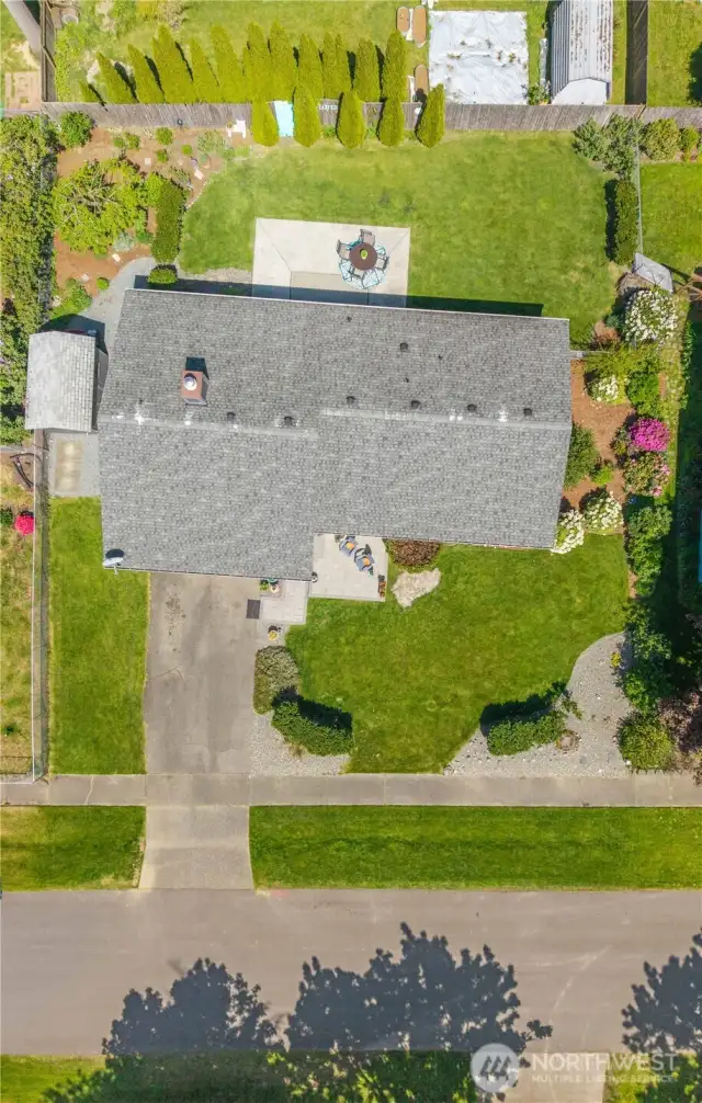 Bird’s-eye view showcasing the expansive, fully landscaped lot with lush green lawn, dual patios (front paver and rear concrete), mature trees, and privacy hedging. This overhead drone shot highlights the layout of the home, backyard entertaining space, garden beds, and overall curb appeal.