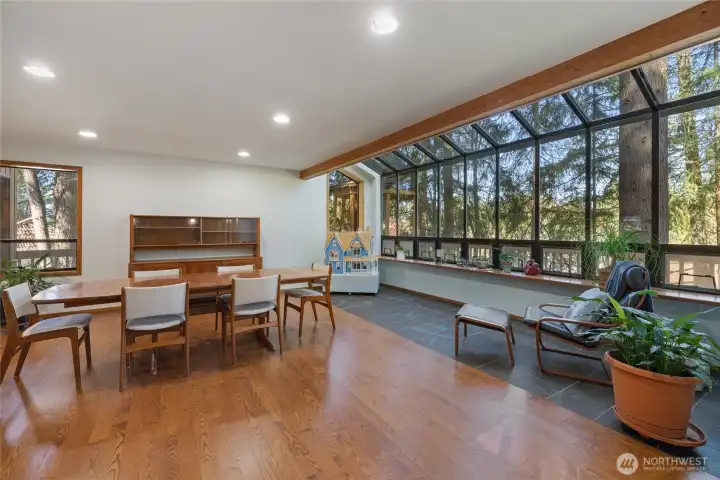 Expansive dining and sitting area with rich hardwood floors and a wall of windows capturing tranquil tree-lined views, creating a bright, inviting atmosphere.