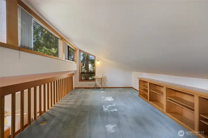 Versatile loft/office space with vaulted ceilings, built-in shelving, and a wall of windows bringing in natural light and treetop views—ideal for work or creative use.
