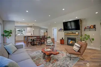 This inviting open-concept living space features a cozy stone fireplace with a mounted TV, stylish modern finishes, and abundant natural light flowing into the updated kitchen—perfect for relaxing or entertaining. Vinyl plank flooring in warm wood tones and designer touches create a welcoming atmosphere you’ll love coming home to.
