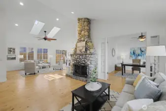 A dramatic vaulted great room anchored by a floor-to-ceiling river rock, wood-burning fireplace creates a warm and inviting centerpiece. Skylights, recessed lighting & walls of windows flood the space with natural light, highlighting the wide-plank hardwood floors. To the right, French doors open to a formal dining room or private office. To the left, the great room flows seamlessly into the family room, kitchen nook & kitchen, offering an ideal layout for both everyday living and entertaining.