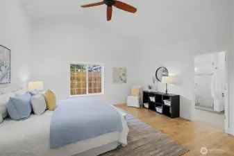 Light-filled primary bedroom showcasing vaulted ceilings, warm wood flooring and a large window that brings in abundant natural light. A ceiling fan adds comfort, while the airy layout and clean architectural lines create a relaxing retreat. The ensuite bath is located just off the bedroom, offering easy access and a seamless flow between spaces.