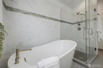 Close-up view of the primary bathroom highlighting the sculptural freestanding soaking tub set against marble-style tile walls with a sleek accent inlay. The adjacent frameless glass shower features modern fixtures and a tiled shower floor for a clean, cohesive look. Soft neutral finishes, crisp white surfaces and contemporary hardware combine to create a polished, tranquil atmosphere with a refined architectural feel.
