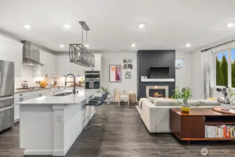 Open concept living space featuring a large white kitchen island with seating, white cabinetry, stainless steel appliances, recessed lighting, and a living area with fireplace and large windows providing natural light.