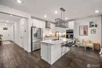 Wide interior view highlighting the open floor plan connecting the white kitchen, living room, and entry hallway with recessed lighting, large center island, stainless steel refrigerator, and built-in wall ovens.
