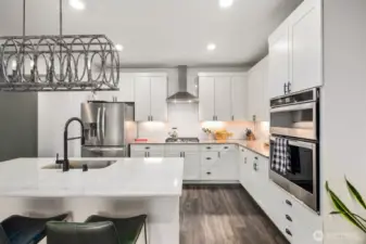 Bright white kitchen workspace with white cabinetry, stainless steel range hood, built-in wall ovens, tile backsplash, and an oversized island with seating positioned under a decorative pendant light fixture.