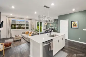Open concept kitchen looking toward the living and dining areas with a large center island, built-in sink, stainless steel dishwasher, recessed lighting, and expansive windows bringing in natural light.