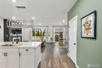Wide interior perspective showing the kitchen island and open living space, with recessed lighting, large windows, and sliding glass door access to the backyard patio.