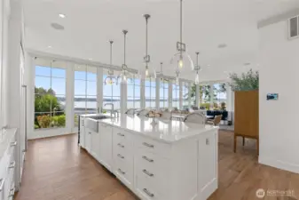 Anchored by quartz counters and custom cabinetry, the kitchen shines with pendant lighting, ample storage, and high-end appliances—Sub-Zero, Bosch, and GE nugget ice maker. From the breakfast counter, the view stretches through the dining room to curated gardens and the water beyond.