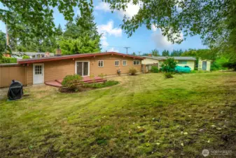 Shady tall trees framing the lawn, grill, and garden space.  Extra storage shed attached on the left.
