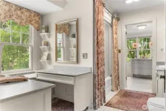 Light-filled upstairs bathroom with charming vintage details, dual vanities, and classic built-in shelving. Timeless design with floral accents, curated shelves, and a private dressing area overlooking the trees.