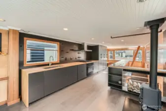 This sleek, modern kitchen stuns with clean lines, hidden appliances, and warm butcher block counters. Matte black cabinetry blends seamlessly with integrated storage, while the gas range and minimalist open shelving complete the contemporary design. A cozy wood stove nearby adds charm and warmth—perfect for life on the lake.