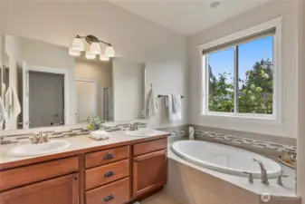 With its soothing palette and thoughtful design, this primary bathroom feels like a personal spa. A deep oval soaking tub is set beneath a large picture window, inviting you to relax while enjoying views of a lush garden. The dual-sink vanity features rich wood cabinetry, a solid-surface countertop, and an eye-catching mosaic tile backsplash that echoes the accent tile around the tub. Abundant natural light, modern fixtures, and a separate shower area (just out of view) complete this bright, elegant space, making it the perfect retreat for both morning routines and evening unwinding.