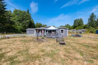 Front exterior of main home with deck, campfire area with a bench, tons of yard space with garden moments throughout, and a 2-car garage with workbench space