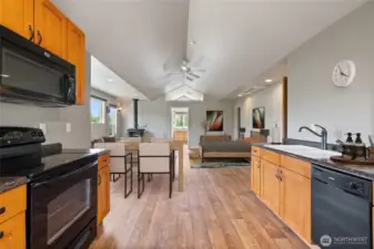 This view highlights the spaciousness of the open-concept living area, featuring vaulted ceilings, a ceiling fan, and a mini-split system. It includes a dining space, full kitchen, and ample room for your creative design ideas. The layout also offers an attached bath, two closets (one walk-in), and additional built-in wall storage for maximum convenience. The free-standing wood stove provides cozy warmth on cool nights.