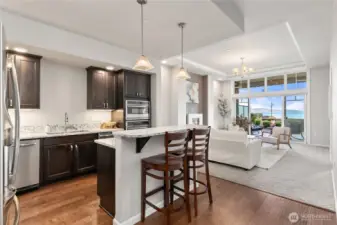 Open-concept kitchen and living space featuring rich cabinetry, granite countertops, stainless steel appliances, and a breakfast bar for casual dining. Pendant lighting and warm wood flooring create an inviting atmosphere that flows seamlessly into the living area and toward expansive waterfront views.