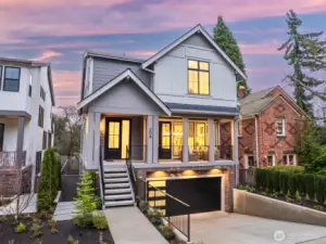 Striking front elevation with a covered porch, warm interior glow, and a drive-under two-car garage that gives this newly built home a polished, timeless presence.