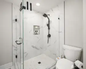 A stunning walk-in shower with full granite slabs on the walls, a niche for soap, dual shower head with a handy wand, tile flooring and glass doors.