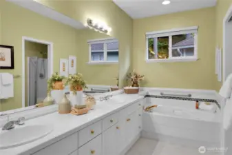 Primary bath off bedroom. Banjo-style tiled countertop at double sink vanity. Tile floor. Paneled white cabinets with shiny brass hardward, brass-accented shiny-chrome faucets. Shiny chrome style light bar with globe lights. Vinyl window with window screen and two-inch blinds. Oval tub with safety hand bar, tile surround and Traditional millwork. Paneled Quaker pocket door to watercloset room with tile floor. American Standard® elongated watercloset. Fiberglass-molded shower with clear glass doors and handheld shower wand. Large walk-in closet with coated ventilated wire organizer shelving.